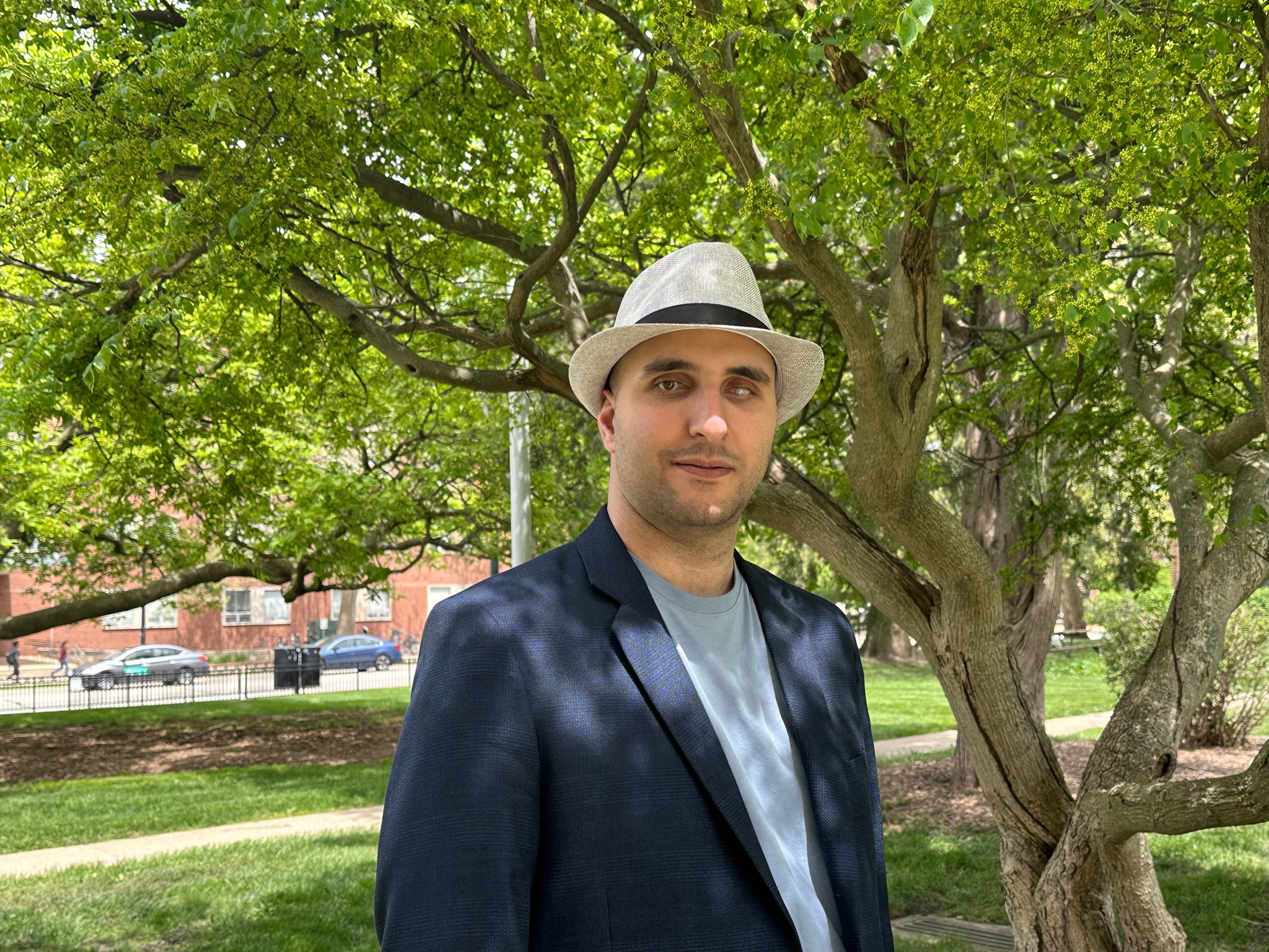 Smiling man wears a light grey fedora, navy blue blazer, and sky blue shirt while standing under a tree.